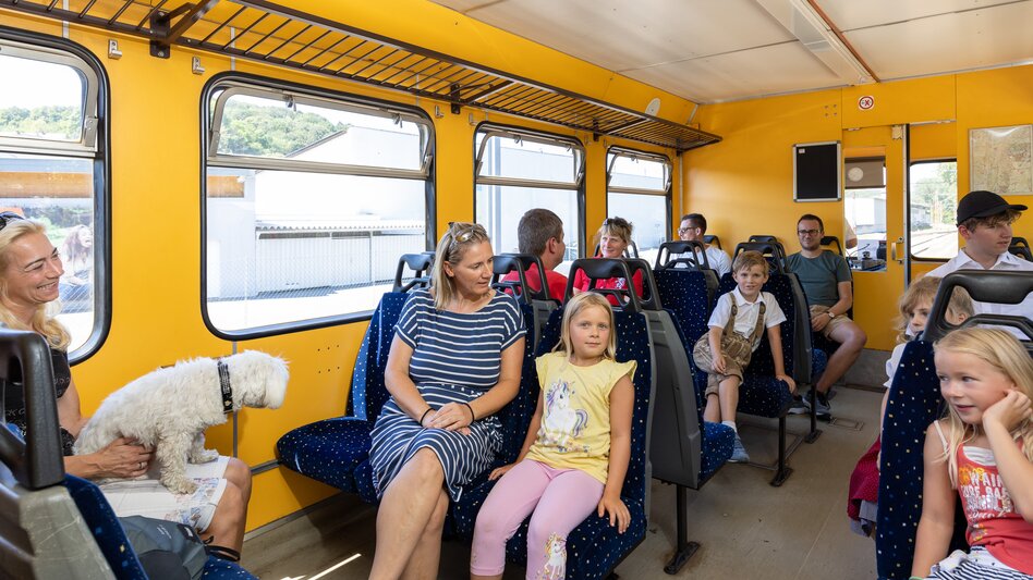 Erwachsene und Kinder sitzen in der Gleichenbergerbahn, deren leuchtend gelbe Innenwände sich von den dunkelblauen Sitzen abheben. Alle freuen sich auf die bevorstehende Fahrt...  | © StB