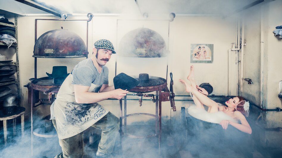 In der nebeligen Hutwerkstatt ist eine alte Badewanne aufgestellt, in der eine Dame badet. Der Hutmacher bietet ihr einen neu angefertigten Hut an. 