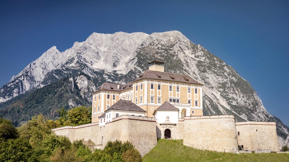 Das helle Schloss Trautenfels erstrahlt gut geschützt von seinen Mauern vor dem anmutigen Grimming, welcher in den wolkenlosen Himmel ragt. | © UMJ