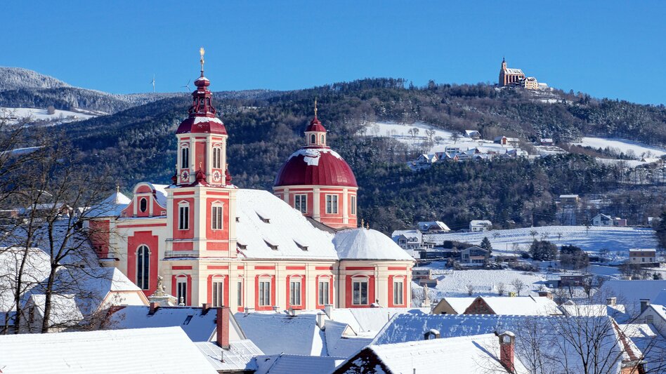 Über die schneebedeckten Dächer vom Pöllauer Zentrum hinweg zeigt das Bild das Schloss Pöllau sowie im Hintergrund die Wallfahrtskirche am Pöllauberg. Es ist alles schneebedeckt bei blauem Himmel und Sonnenschein.