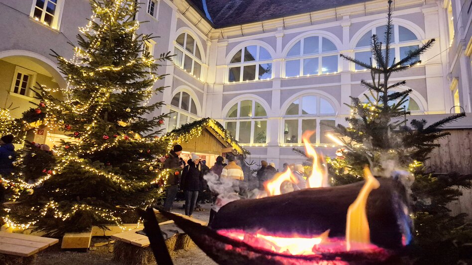 Der Innenhof im Schloss Pöllau ist mit Kerzen und Lichterketten erhellt, christbäume schmücken den Hof und Stände mit Kulinarik und Handwerk laden zum vorweihnachtlichen Genuss ein. Im Vordergrund ist auch eine Feuerschale mit brennenden Holzscheitern zu sehen.