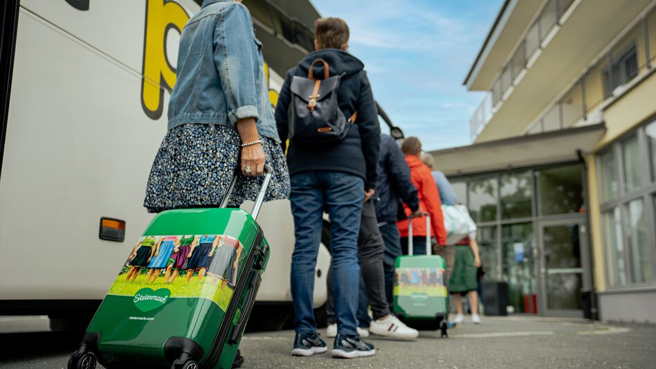 Eine Gruppe von Gästen mit Steiermark-Koffer neben Bus und vor dem Hotel-Eingang