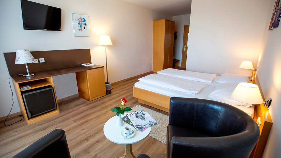 Das Doppelzimmer Comfort im Hotel Novapark ist mit Holzboden und Holzmöbeln in Brauntönen eingerichtet. Auch ein Schreibtisch und eine Sitzecke sind vorhanden. TV und Stehlampe sind zu sehen.