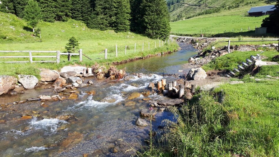 ein Bach umgeben von saftig grünen Almwiesen inmitten eines Tales, dahinter Berge und Weiden