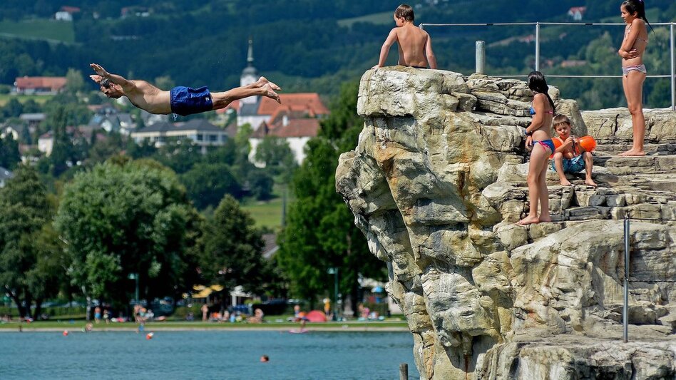 Ein Mann springt von einem Sprungfelsen ins Wasser. Kinder beobachten ihn dabei. Im Hintergrund ist der Stubenbergsee  zu sehen.