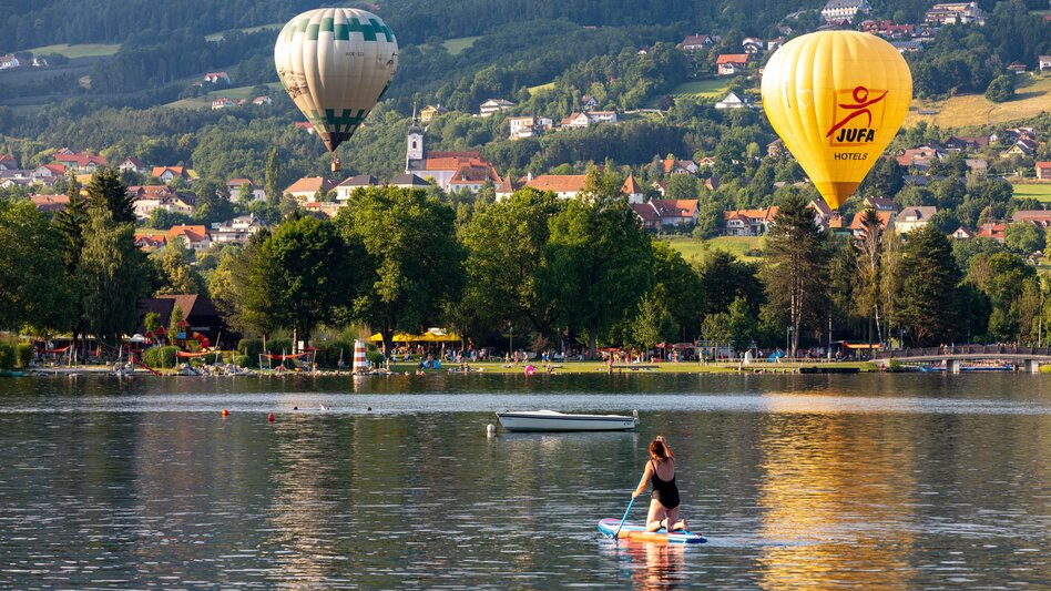 Eine Frau fährt mit dem Stand-Up-Paddle über den Stubenbergsee. Im Hintergrund sind groß zwei Heißluftballone zu sehen. | © Gemeinde Stubenberg am See