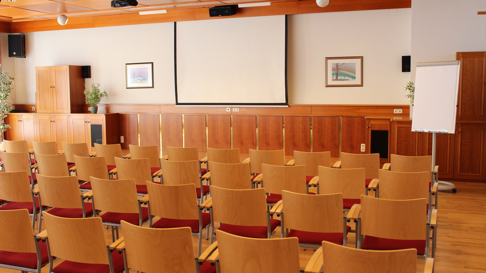 Im Mehrzweckraum sind Sesselreihen aufgebaut. Vorne steht ein Flipchart, die Leinwand für den Projektor ist ausgerollt.