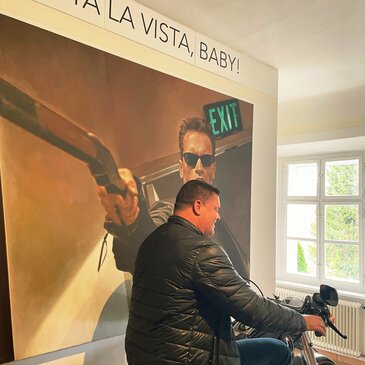 Beeindruckendes Arnold Schwarzenegger Museum. Ein Teilnehmer sitzt hier am Motorrad und im Hintergrund ist ein großes Poster an der Wand mit Arnold Schwarzenegger und dem Schriftzug "Hasta la vista, Baby!" zu sehen.