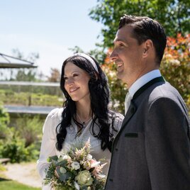 Brautpaar bei einer freien Trauung im Garten des Retter Bio-Natur-Resorts an einem sonnigen Tag. Die Braut hält einen Blumenstrauß, beide lächeln und stehen inmitten einer natürlichen, grünen Umgebung.
