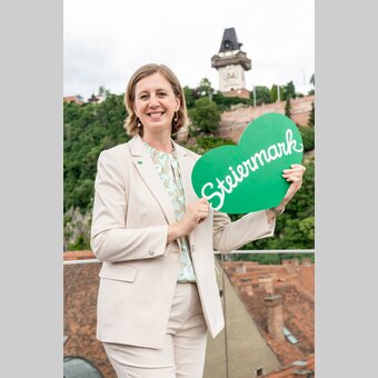 Tourismuslandesrätin Barbara Eibinger-Miedl mit dem Grünen Herz, im Hintergrund der Grazer Schlossberg als beliebtestes Ausflugsziel.  | © STG | Jesse Streibl