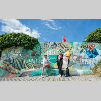 Künstler Tom Lohner und Zur Schmied´n-Gastgeber Anna-Lisa und Toni Maier mit der neuen Wandermalerei beim Gasthaus Zur Schmied´n.  | © STG | Jaden Gynes