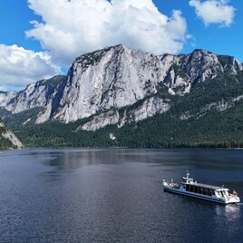 Das Solarschiff der Schifffahrt Altaussee gleitet an einem schönen Sommertag über den malerischen Altausseer See. Im Hintergrund ist die eindrucksvolle Trisselwand unter blauem Himmel zu sehen. | © Schifffahrt Altaussee