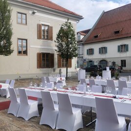 A festively set table outdoors with white chairs and red decorations. In the background, historic buildings and trees can be seen.