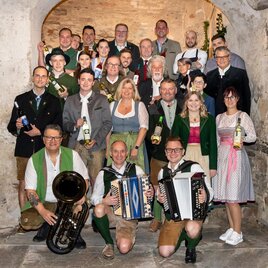 A group of people in traditional clothing, smiling and holding various drinks. They are standing in a historical room with stones and warm lighting. | © Fotostudio Augenblick