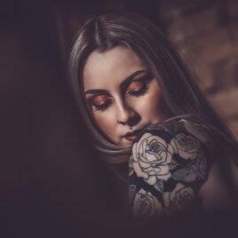 Eine Frau mit langen, grauen Haaren und auffälligem Make-up. Ihr Arm ist mit einem kunstvollen Tattoo von Rosen verziert.