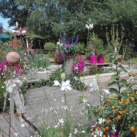 Ein malerischer Garten mit bunten Blumen und Pflanzen. Holzstege und blühende Töpfe verleihen der Szene eine einladende Atmosphäre. | © Manuela Schweinzer-Riegler