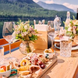 <p>Ein schön gedeckter Tisch mit Köstlichkeiten und Blumenarrangements am Wasser. Im Hintergrund sind Berge und ein klarer Himmel zu sehen.</p> | © Tauroa