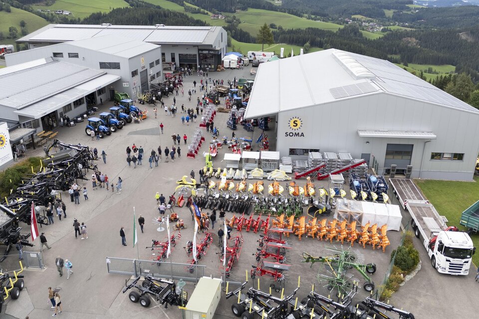 Ein großer Betriebshof mit vielen Besuchern und landwirtschaftlichen Maschinen ist zu sehen. Im Hintergrund gibt es grüne Hügel und Gebäude. | © SOMA