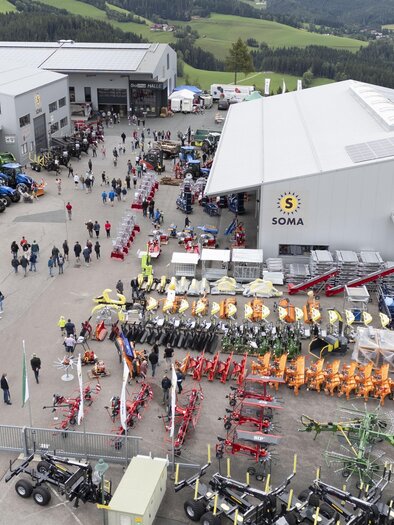 Ein großer Betriebshof mit vielen Besuchern und landwirtschaftlichen Maschinen ist zu sehen. Im Hintergrund gibt es grüne Hügel und Gebäude. | © SOMA