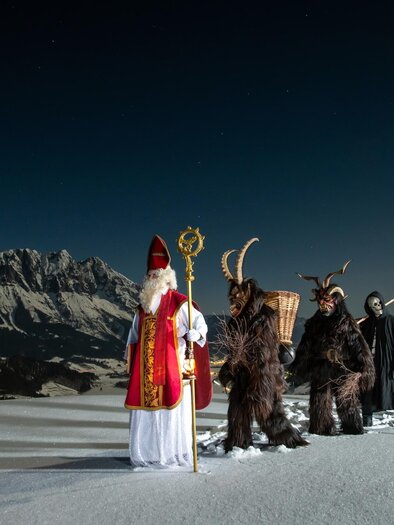 Eine winterliche Landschaft mit schneebedeckten Bergen. Im Vordergrund stehen Figuren in traditionellen Kostümen, darunter ein Heilige mit einem Stab und mystische Begleiter. | © Öblaner Krampusgruppe / Christoph Huber