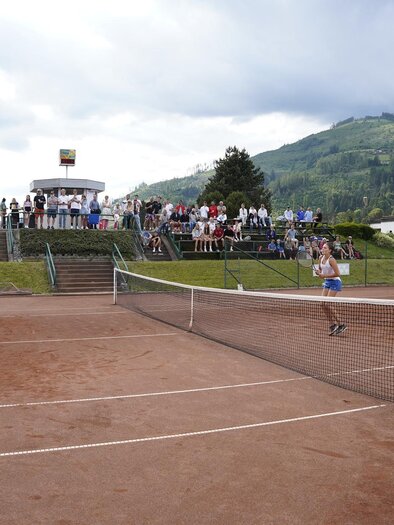 Two female players are playing tennis on a clay court. A group of spectators is watching the game in the background. | © Stadt Bruck