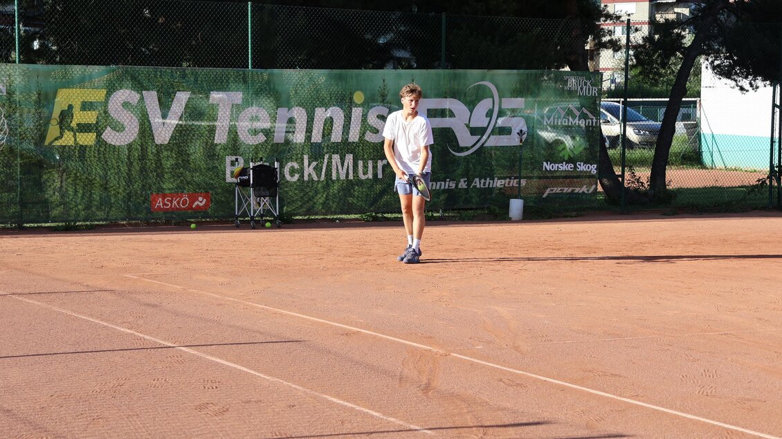 Ein Tennisplatz mit rotem Sand und einem Spieler, der sich auf den Aufschlag vorbereitet. Im Hintergrund ist ein Werbebanner des Tennisvereins sichtbar. | © Stadt Bruck