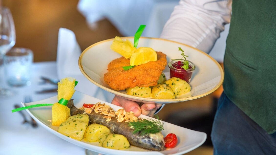 Ein Kellner serviert zwei Teller mit köstlichen Gerichten. Oben ein paniertes Schnitzel mit Kartoffeln und unten ein gebackener Fisch mit Kräutern und Beilagen. | © Hotel Seevilla Altaussee