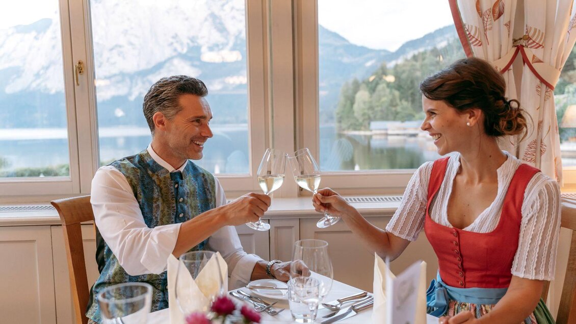 Ein Paar stößt mit Gläsern an, während sie an einem schön gedeckten Tisch sitzen. Im Hintergrund sind Berge und ein ruhiger See zu sehen. | © Karl Steinegger