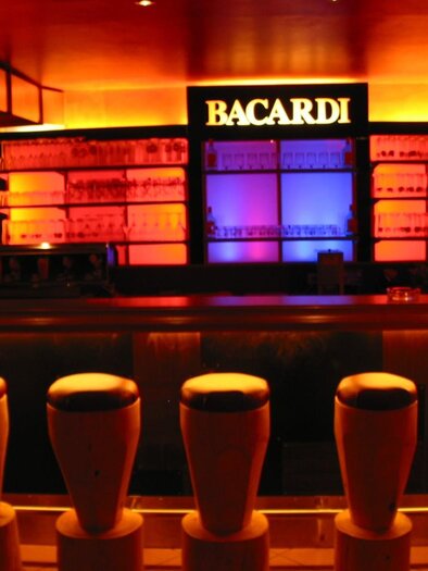 A stylish bar with illuminated shelves and colorful lights. The bar stools are arranged in a modern and inviting way. | © Hotel Seebacherhof