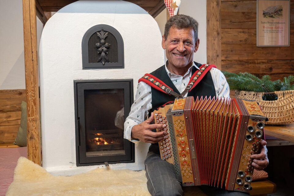 Ein lächelnder Mann in traditioneller Tracht mit rotem Oberteil und grünen Details. Er zeigt mit einer Hand auf eine unsichtbare Szene.