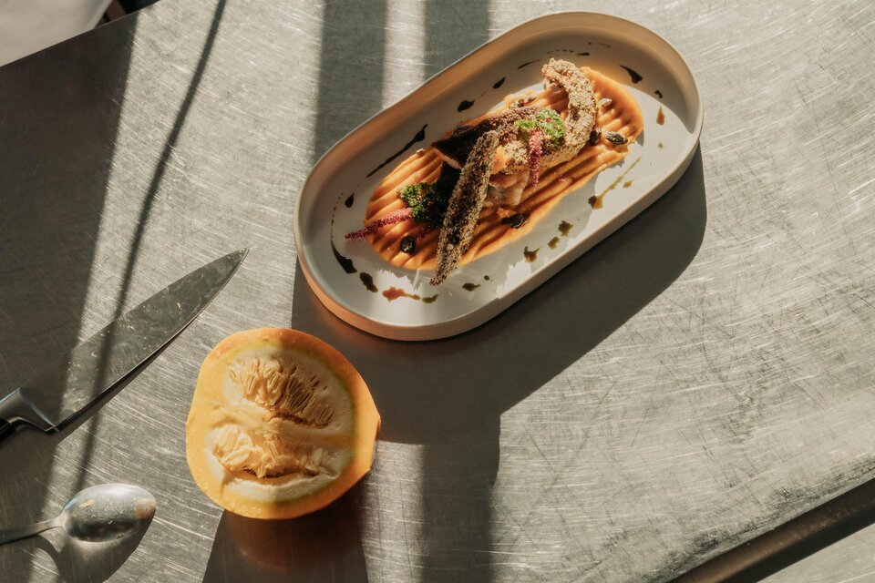 Ein stilvolles Gericht auf einem Teller mit Fisch und Sauce. Neben dem Teller liegt eine halbierte Frucht. | © studio-draussen