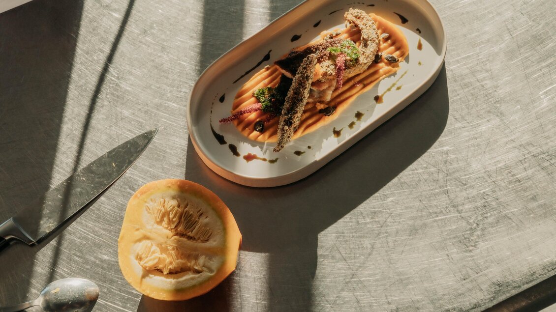 Ein stilvolles Gericht auf einem Teller mit Fisch und Sauce. Neben dem Teller liegt eine halbierte Frucht. | © studio-draussen