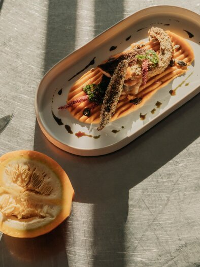 Ein stilvolles Gericht auf einem Teller mit Fisch und Sauce. Neben dem Teller liegt eine halbierte Frucht. | © studio-draussen