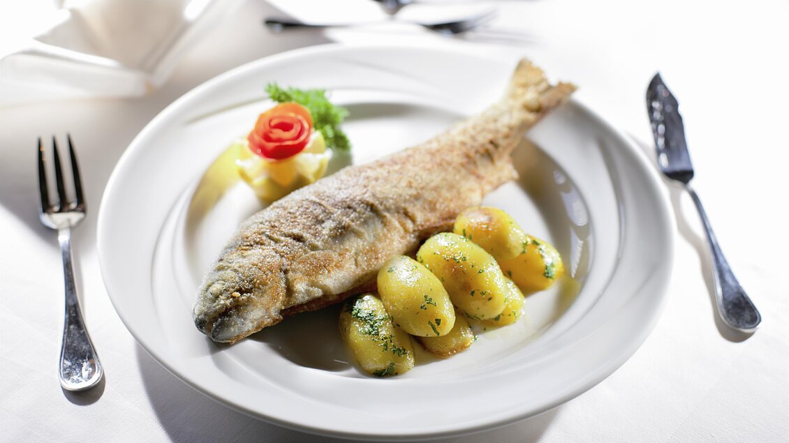 Ein gebratener Fisch liegt auf einem Teller mit Kartoffeln und einer Garnitur aus Gemüse. Die Tischdecke ist hell und das Besteck ist ordentlich arrangiert. | © GH Donner