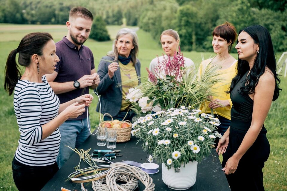 Eine Gruppe von sieben Personen trifft sich im Freien, um gemeinsam mit Blumen und Pflanzen zu arbeiten. Auf einem Tisch liegen Materialien und frische Blumen zum Basteln bereit. | © Karin Heschl-Polzhofer