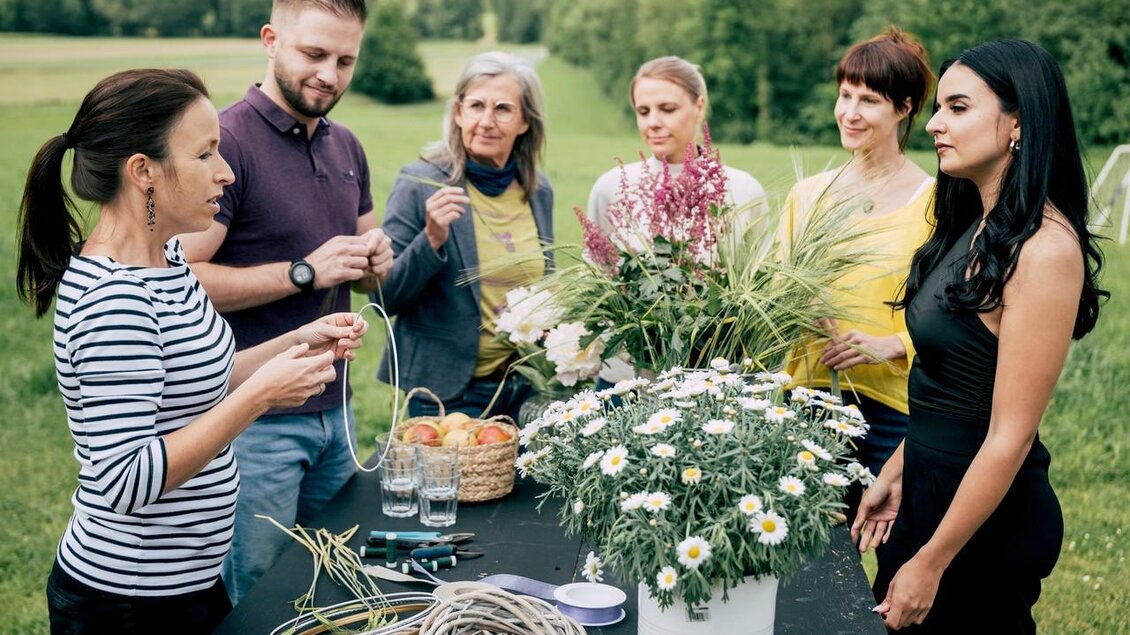 Eine Gruppe von sieben Personen trifft sich im Freien, um gemeinsam mit Blumen und Pflanzen zu arbeiten. Auf einem Tisch liegen Materialien und frische Blumen zum Basteln bereit. | © Karin Heschl-Polzhofer