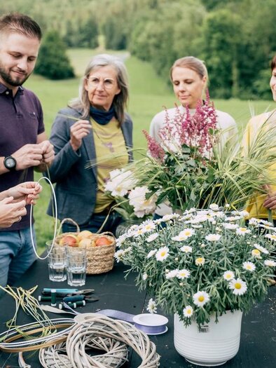 Eine Gruppe von sieben Personen trifft sich im Freien, um gemeinsam mit Blumen und Pflanzen zu arbeiten. Auf einem Tisch liegen Materialien und frische Blumen zum Basteln bereit. | © Karin Heschl-Polzhofer