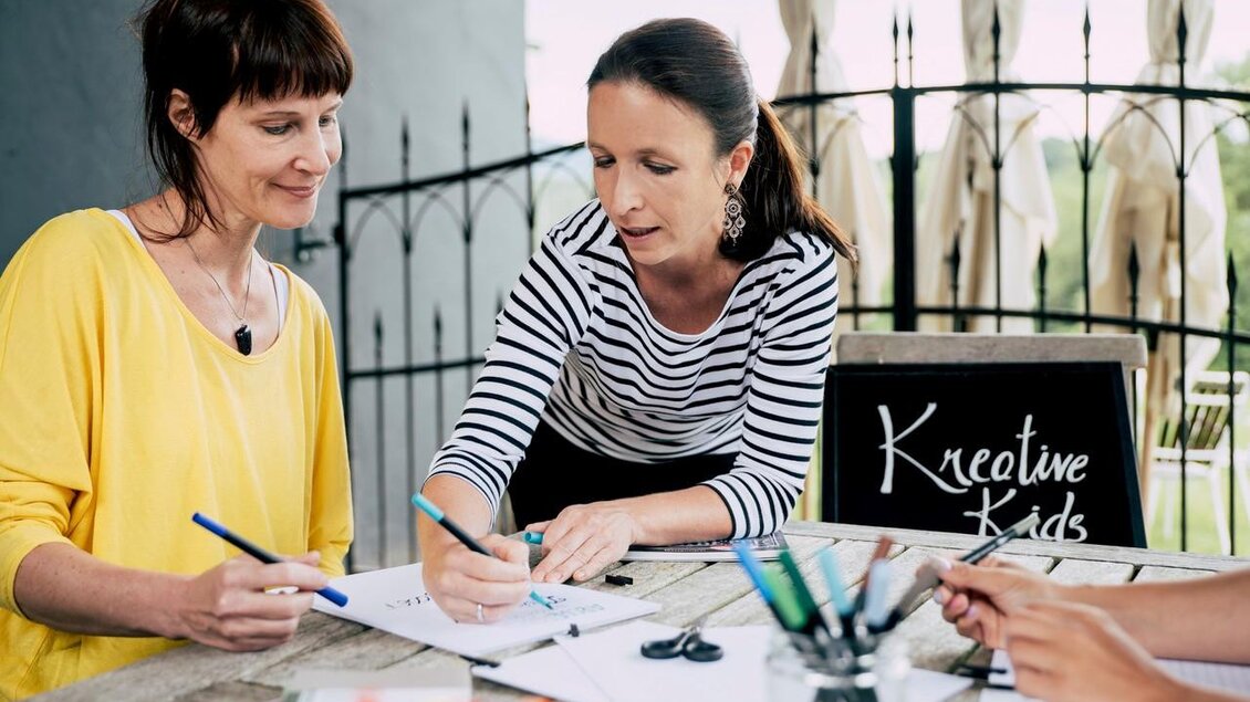 Zwei Frauen arbeiten an einem Tisch und zeichnen mit Stiften. Im Hintergrund steht ein Schild mit der Aufschrift "Kreative Kids". | © Karin Heschl-Polzhofer