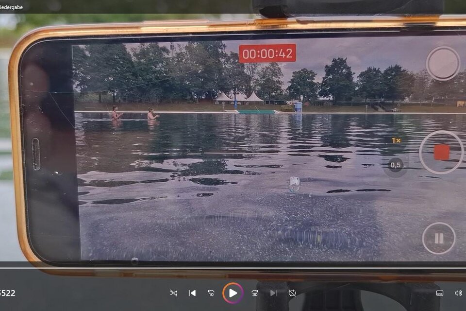 Ein Smartphone filmt die ruhige Wasseroberfläche eines Naturbades und hält die entspannte Atmosphäre aus einer ungewöhnlichen Perspektive fest. Im Hintergrund sind Badestege, Schwimmende und die weitläufige Parklandschaft zu sehen. Zu sehen bei der Wasser Biennale in Fürstenfeld. | © raumarbeiterinnen