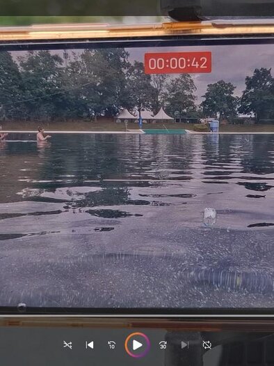 A smartphone films the calm water surface of a natural swimming pool and captures the relaxed atmosphere from an unusual perspective. In the background, there are swimming docks, swimmers, and the expansive park landscape. On display at the Water Biennale in Fürstenfeld. | © raumarbeiterinnen