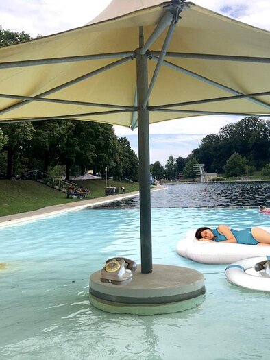 A generous natural pool combines crystal clear water with a relaxed atmosphere of well-being. Shady sun umbrellas, cozy swimming and relaxation areas, as well as the spacious park landscape invite you to unwind and enjoy on warm summer days. Art installation at the outdoor pool Fürstenfeld. | © Glassner&Serasini