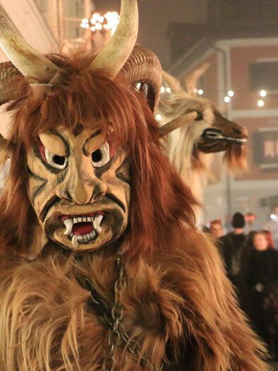 A crowd is celebrating a traditional event. In the foreground stands a person in an impressive costume with a masked face and animal elements. | © Silvia Rastl