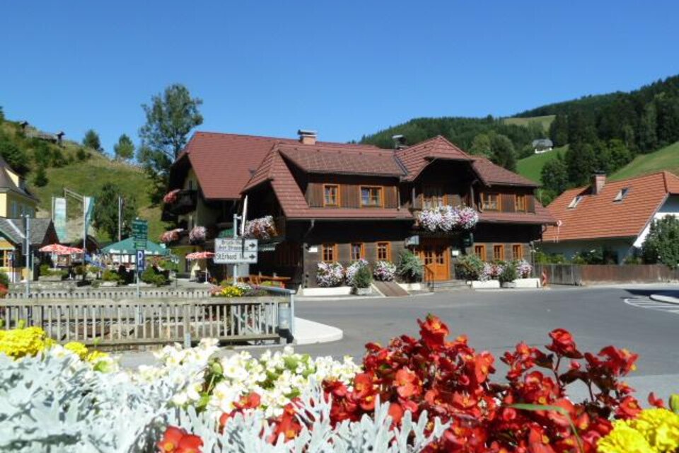 Ein charmantes Holzhaus mit bunten Blumenkästen und einer malerischen Umgebung. Im Vordergrund blühen farbige Blumen, während die Landschaft im Hintergrund sanfte Hügel zeigt. | © Gasthof Grabenbauer