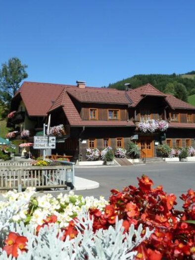 Ein charmantes Holzhaus mit bunten Blumenkästen und einer malerischen Umgebung. Im Vordergrund blühen farbige Blumen, während die Landschaft im Hintergrund sanfte Hügel zeigt. | © Gasthof Grabenbauer