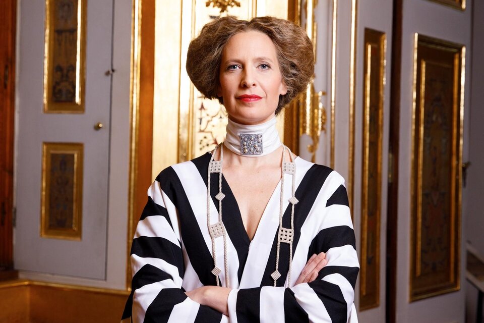 Porträt einer elegant gekleideten Frau in einem auffälligen schwarz-weiß gestreiften Gewand. Mit verschränkten Armen steht sie selbstbewusst vor einer prachtvollen, goldverzierten historischen Innenausstattung. Das Bild verbindet moderne Eleganz mit einem stilvollen, herrschaftlichen Ambiente. Maxi Blaha im Zentrum Feldbach. | © Peter Rigaud