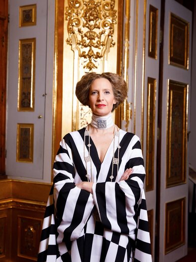Porträt einer elegant gekleideten Frau in einem auffälligen schwarz-weiß gestreiften Gewand. Mit verschränkten Armen steht sie selbstbewusst vor einer prachtvollen, goldverzierten historischen Innenausstattung. Das Bild verbindet moderne Eleganz mit einem stilvollen, herrschaftlichen Ambiente. Maxi Blaha im Zentrum Feldbach. | © Peter Rigaud
