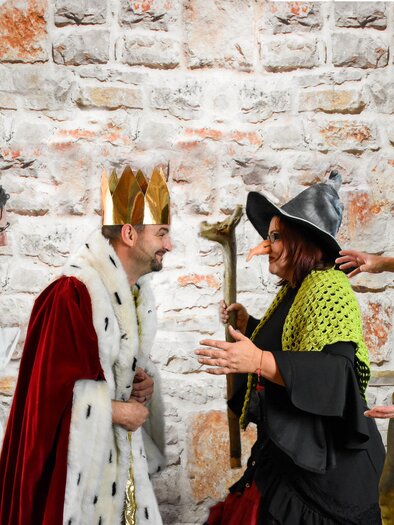 Eine fröhliche Theatergruppe in fantasievollen Kostümen – darunter König, Hexe und weitere Märchenfiguren – steht vor einer rustikalen Ziegelwand. Die lebendige Szene vermittelt Humor, Spielfreude und beste Unterhaltung für die ganze Familie im Zentrum Feldbach. | © SET