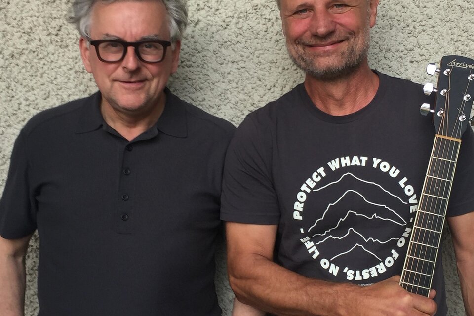 Zwei Musiker stehen entspannt nebeneinander vor einer schlichten Wand. Einer der beiden hält eine Akustikgitarre in der Hand. Das sympathische, ungezwungene Porträt vermittelt Nähe und Vorfreude auf einen musikalischen Abend im Zentrum Feldbach. | © Rudi Ferder