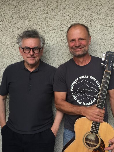 Zwei Musiker stehen entspannt nebeneinander vor einer schlichten Wand. Einer der beiden hält eine Akustikgitarre in der Hand. Das sympathische, ungezwungene Porträt vermittelt Nähe und Vorfreude auf einen musikalischen Abend im Zentrum Feldbach. | © Rudi Ferder
