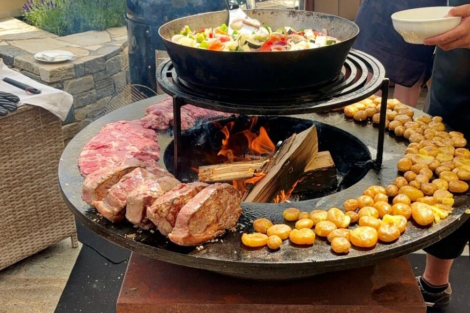 Ein Grill mit verschiedenen Fleischsorten und Gemüse wird zubereitet. Im Hintergrund sind grüne Hügel und ein klarer Himmel zu sehen. | © Tourismusverband Oststeiermark